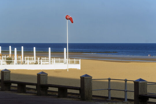 A tall mast topped with a windsock rises above the sand of Ostend, facing the calm sea and clear sky, near the beach fence.