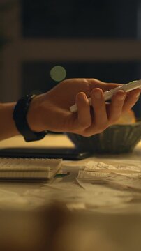 Young adult sits at a dimly lit table, writing notes on paper while glancing at a phone surrounded by scattered receipts, creating a focused late-night setting for personal finance management