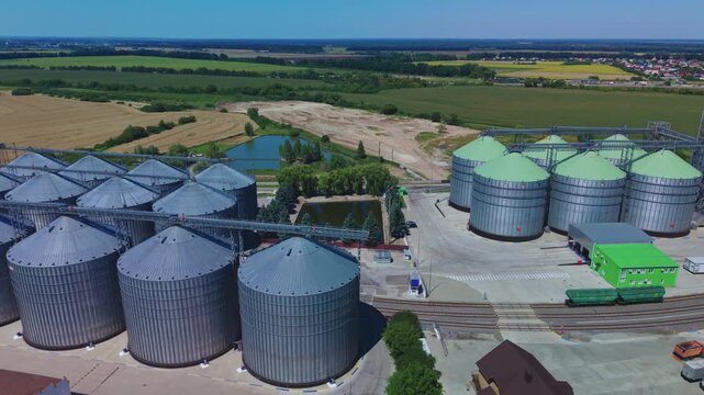 Dynamic aerial overview reveals rows of silver and green metal silos at modern grain storage terminal with railway tracks and cargo cars.