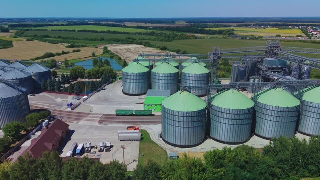 Dynamic aerial overview shows large silver and green metal silos of modern grain elevator terminal near countryside fields and railway lines.