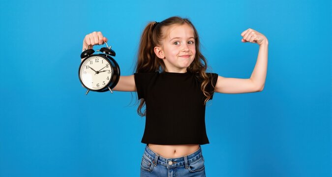 Young girl wearing black crop top and denim jeans holds alarm clock while stretching her arm against a solid blue background in a playful pose
