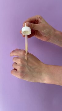 Golden serum drops from a cosmetic dropper onto hands, then hands rub rubbing serum together on purple background. Skincare application, beauty swatches, facial serum, cosmetic product, UGC video