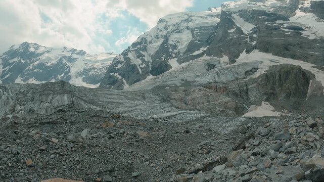 Rugged alpine landscape featuring snow capped peaks, glacier formations, rocky foreground under partly cloudy sky. Cool tones dominate with gray rock, white snow, blue sky