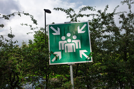 A green muster point sign stands on a pole beside a street lamp at a military training area.