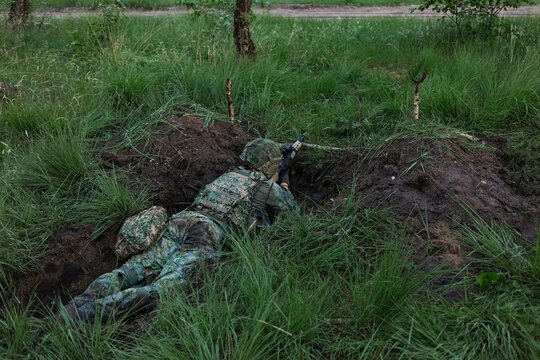 A soldier aims a service rifle from a prone fighting position in tall grass during a military exercise.