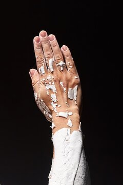 Close-up of a hand covered in white clay cracking and peeling to reveal skin against a black background