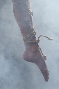 Woman's leg and foot bound with intricate shibari ropes against a smoky, gray studio background.