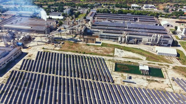 Solar panels are placed in rows on the ground near factories. The scene shows smokestacks and a body of water. This view highlights the use of renewable energy in an industrial area.