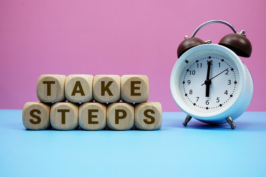 Take Steps alphabet letters wooden cube with alarm clock on pink and blue background
