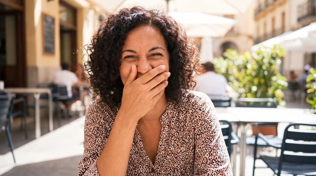 A happy woman with curly hair laughs heartily, covering her mouth with her hand while sitting at an outdoor cafe on a sunny day.