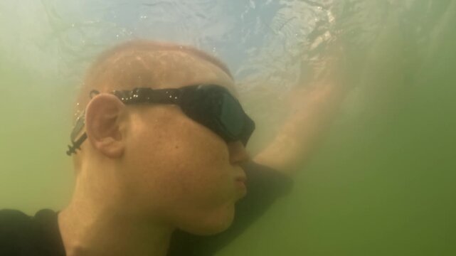A teenager wearing swimming goggles takes a selfie with an underwater camera. A boy dives with a camera to take a photo