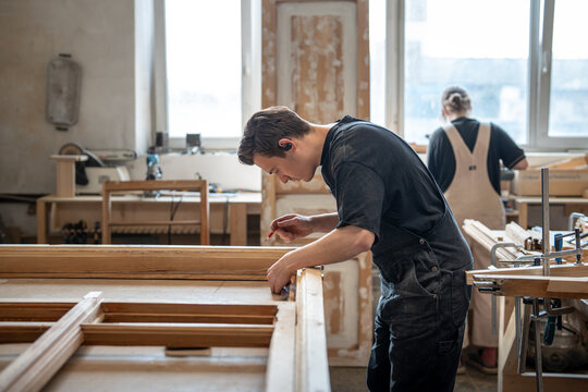 Concentrated craftsman working with antique wooden window frame at workplace in renovation studio, thoughtful man carpenter measuring details and parts of vintage frame using ruler and pencil tools.