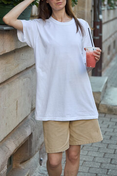 Woman in white t-shirt and beige shorts holding a pink drink outdoors