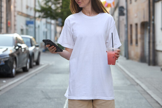 Woman in white t-shirt holding a pink drink and phone on a city street