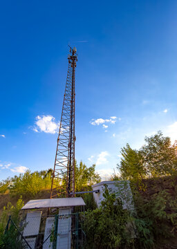 Cell tower stands tall in clear sky. A cell tower is visible against the blue sky with clouds. Green trees surround the base of the tower in the area.