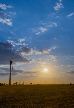 Sunset over a field with wind turbine. A wind turbine stands tall in a field as the sun sets in the background. Clouds fill the sky during the evening.