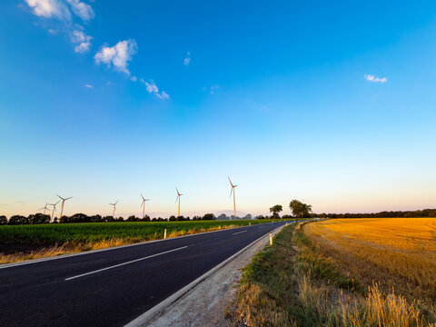 Winding road by turbines at dusk. A road runs along a field with wind turbines in the distance during the evening.