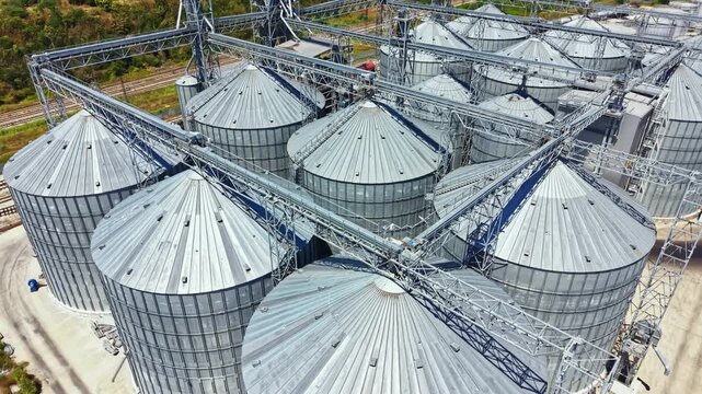 A large farm features multiple grain silos arranged in rows. Structures are present for handling grain. The area is sunny and close to train tracks.