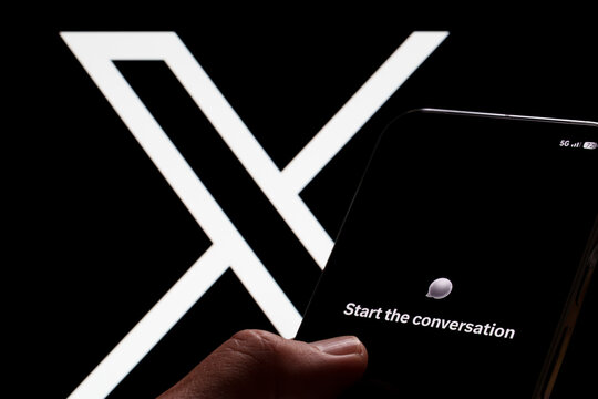 XChat welcome screen displayed on a smartphone held in hand with a large X logo on a monitor behind it. Stafford, United Kingdom, August 2, 2026.
