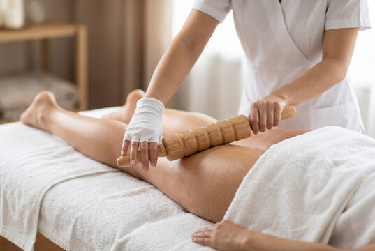 Therapist performing maderotherapy massage on female thigh with wooden roller