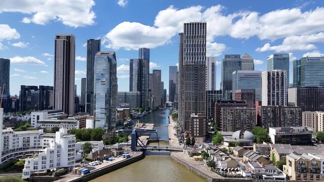 Aerial push-in toward Canary Wharf financial district and West India Dock, showcasing modern skyscrapers, waterfront architecture and the iconic London Docklands skyline beneath a bright blue summer s