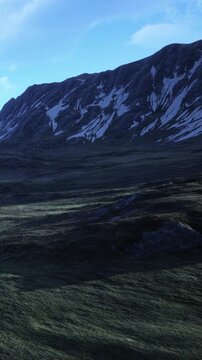 A large mountain stands tall with patches of snow. The landscape is grassy and rugged with a clear blue sky above. Sunlight highlights the mountains features during the day.