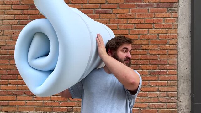 4K video. 60 fps. Mature man with beard stands near house brick wall, holds blue bundle of foam rubber for cheap furniture and looks back. Concept of rent apartment, repair work, dream, sleep
