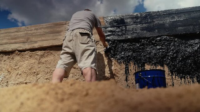 Professional construction worker applying a protective layer of black mastic waterproofing to a building's concrete foundation wall, using a brush for insulation and damp proofing