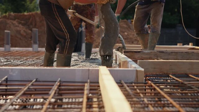 Professional builders pouring liquid concrete into the reinforcing steel mesh of a house foundation slab, using a concrete pump hose and vibrating tool to create a solid and durable structure