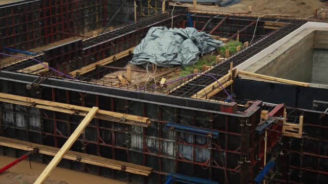 Heavy raindrops falling into puddles of muddy water at a house foundation construction site, showing formwork panels, rebar reinforcement cages, and a recently poured concrete basement wall