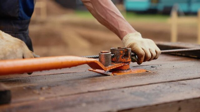 Unrecognizable worker in protective gloves manually bending a steel reinforcing bar using a special machine tool on a wooden table at a construction site, demonstrating physical labor