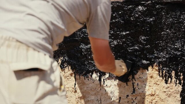 Professional construction worker using a brush to apply black waterproofing tar sealant to a building's concrete foundation wall, protecting the structure from moisture and water damage