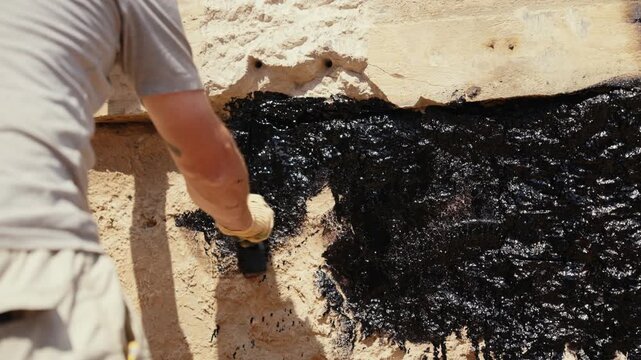 Professional builder applying a thick, black bituminous mastic with a brush to waterproof the foundation of a building, protecting the structure from water damage during renovation