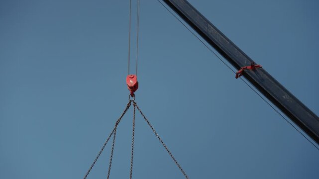 Close up of a mobile crane's red hook and metal chains lifting a heavy load, with the boom arm moving slowly against a clear blue sky during a construction or engineering project