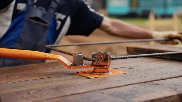 Unidentifiable male worker in protective gloves using a manual bending machine to shape a steel reinforcement bar on a wooden surface at a residential construction site during the day