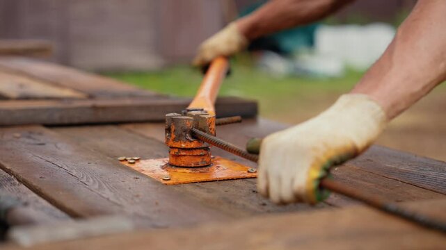 Professional builder in protective gloves bending a metal reinforcement bar with a manual bender tool on a wooden surface at a construction site, demonstrating physical labor and building skills