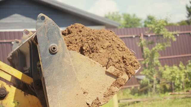 Heavy machinery at a construction site with a front loader moving earth for landscaping. The yellow bulldozer scoop is full of dirt, operating outdoors in a backyard during a sunny day