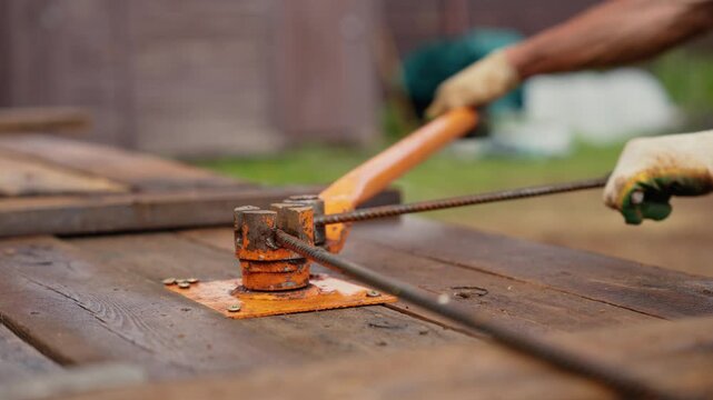 Strong construction worker in protective gloves using a manual rebar bending tool on a wooden surface to shape a steel bar for a reinforced concrete structure at a building site