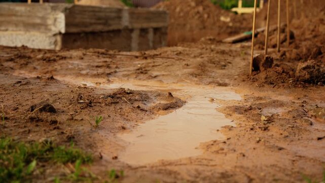 Raindrops falling into a muddy puddle on the ground at a construction site during a downpour. Wet earth and a concrete foundation are visible in the background, creating a damp, earthy scene