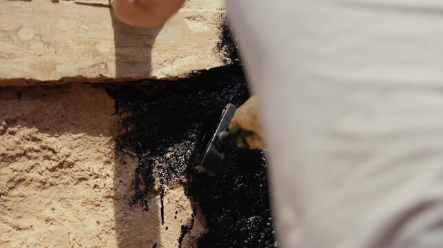 Close up view of a professional worker applying black waterproofing mastic to a concrete foundation wall with a brush, ensuring damp proof protection for the building during construction