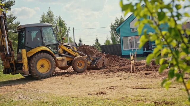 Heavy machinery performing earthmoving work as a yellow backhoe loader scoops up soil with its front bucket, preparing the land for building and leveling the ground near a suburban house