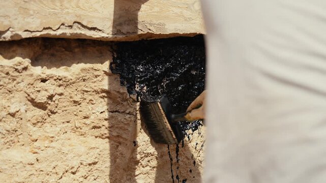 Professional builder applying black bitumen tar with a large brush to a stone foundation wall for waterproofing, sealing joints and protecting the structure from moisture and water damage