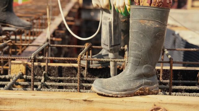 Male builder in rubber boots using a handheld concrete vibrator to settle wet cement inside a rebar cage during foundation work, ensuring structural integrity of the building