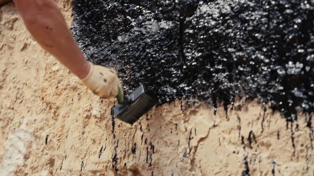 Close up view of a professional builder's hand applying black mastic with a brush for foundation waterproofing, ensuring robust protection and insulation against water damage on a concrete wall