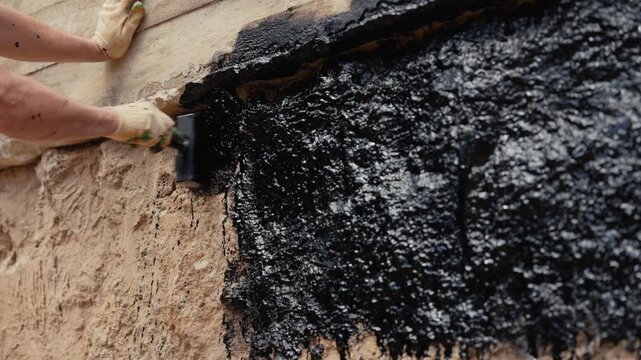 Close up shot of a builder's hands in gloves applying a thick black waterproofing mastic with a brush to a concrete foundation wall, ensuring protection against moisture and water damage