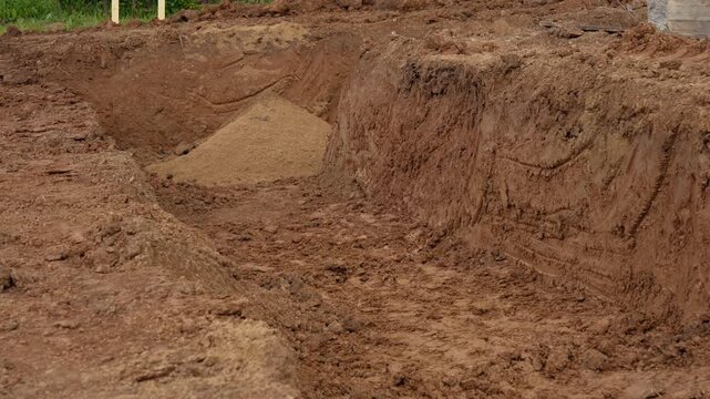 Large excavator bucket digging a deep trench in the ground for a building foundation, with a pile of sand visible at the bottom of the pit during earthworks on a construction site