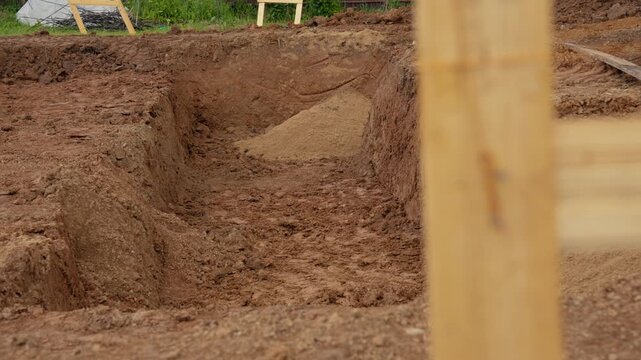 Deep trench dug in the earth for a building foundation at a new construction site. A pile of sand for the base layer is visible at the bottom of the pit, prepared for construction work