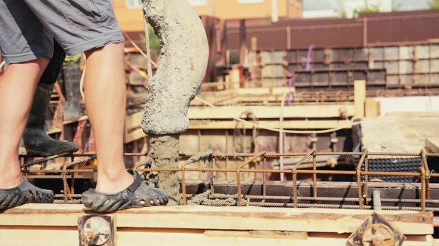 Team of builders pouring wet cement mixture from a pump truck hose into a reinforced formwork with steel rebar on a sunny day. The process of creating a solid house foundation at a worksite