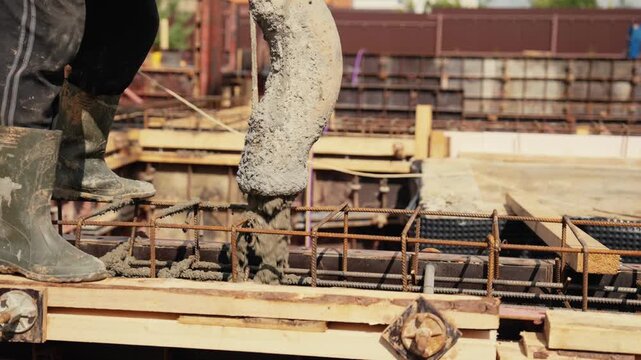 Close up of a worker in rubber boots supervising the process of pouring liquid concrete from a pump hose into a reinforced formwork to create a solid building foundation at a construction site