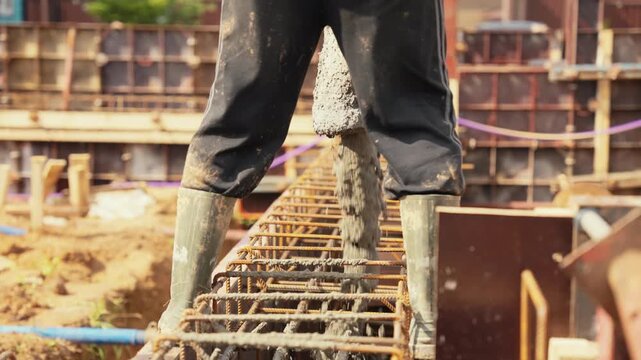 Anonymous builder in work boots pouring wet cement from a pump hose into a rebar cage, creating a reinforced concrete foundation for a new building on a construction site during a sunny day